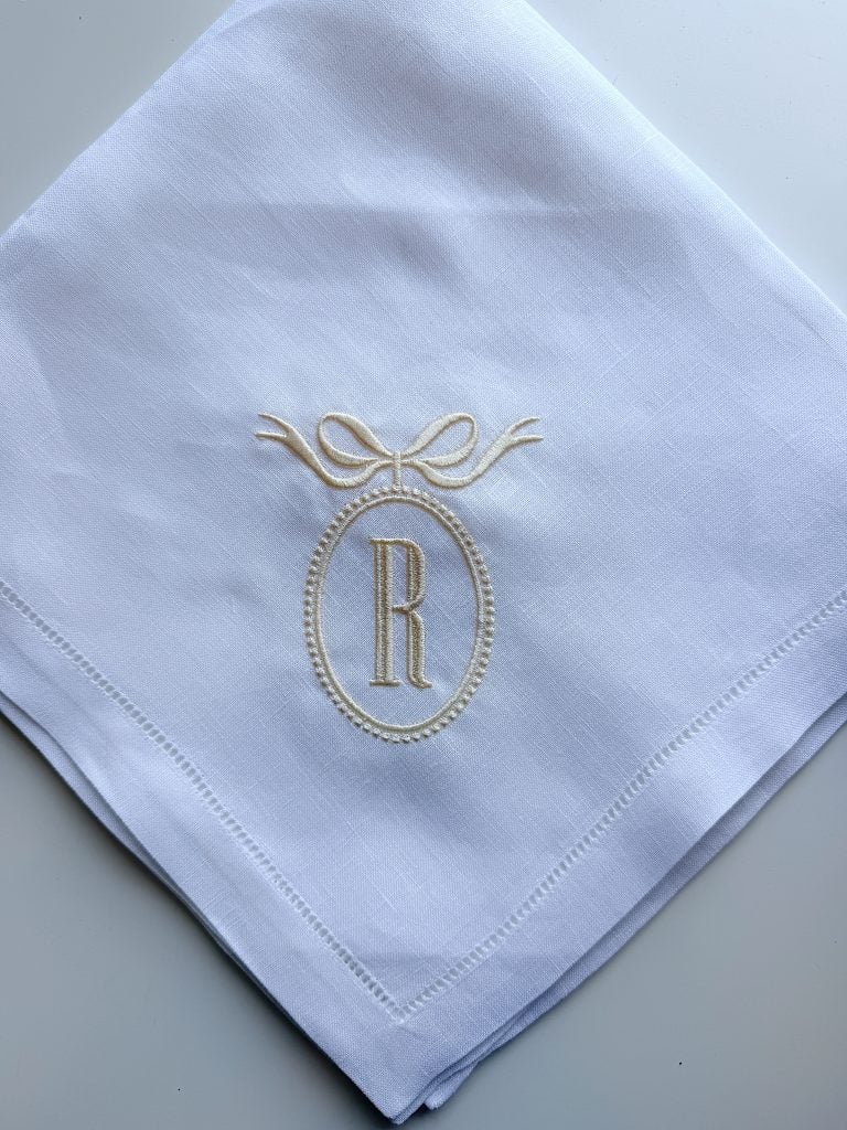 White handkerchief with an embroidered design featuring the letter "R" inside an oval frame topped with a bow, placed on a plain surface.