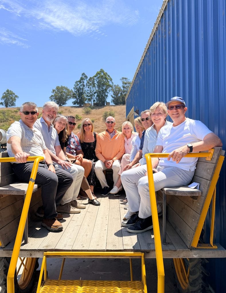A group of twelve adults sit together on a wooden wagon with yellow rails, parked outdoors on a sunny day next to a blue metal wall.