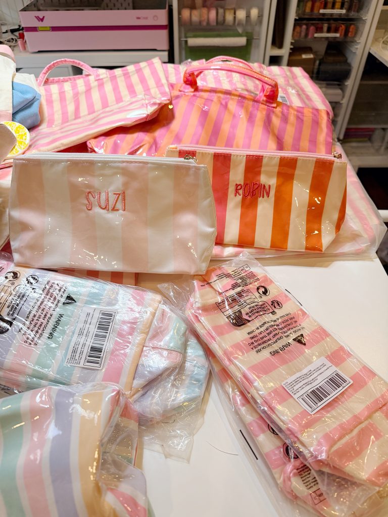 Several striped toiletry bags, some with names "SUZI" and "ROBIN," are displayed on a table, with pastel-striped items in plastic packaging in the foreground.