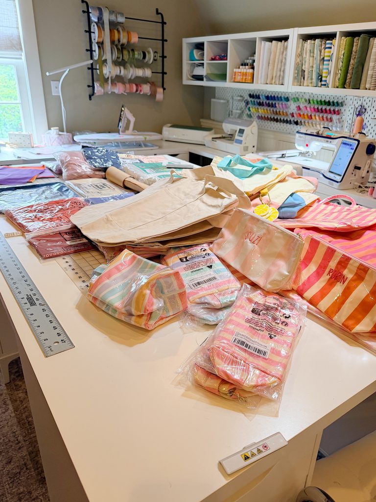 A crafting workspace with sewing machines, rulers, organized thread spools, fabric, tote bags, and packaged fabric items arranged on a large white table.