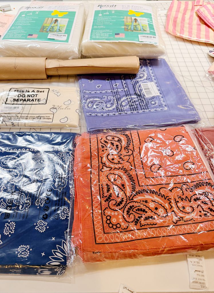 Several folded bandanas in blue, orange, and purple, and packaged sewing materials are arranged on a table with a gridded cutting mat.