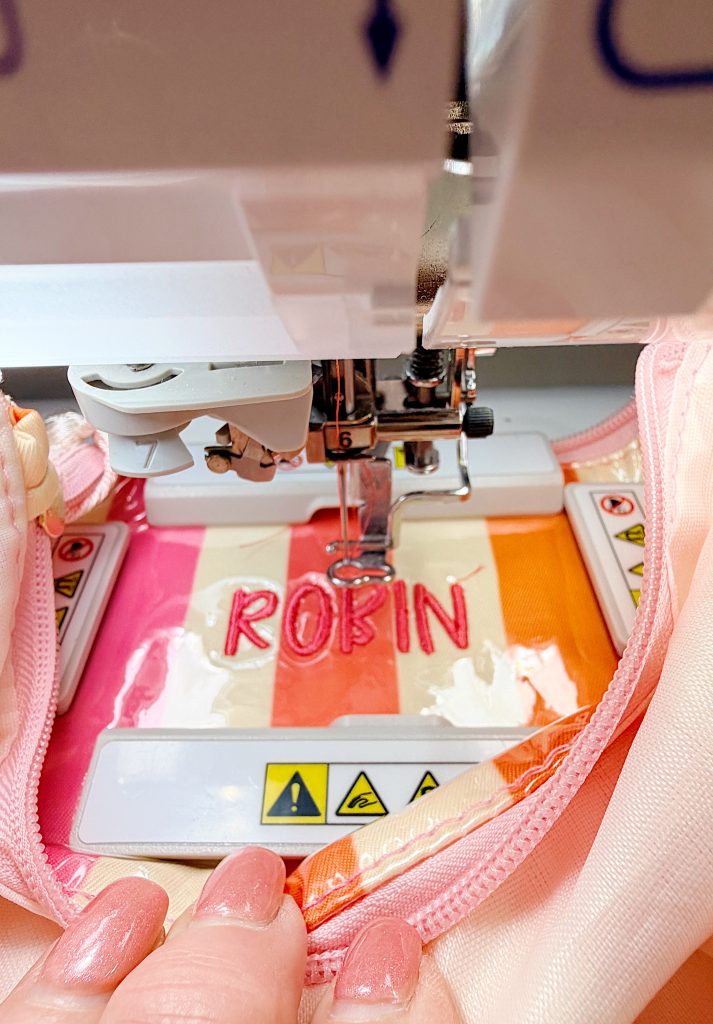 A sewing machine embroiders the name "ROBIN" in pink thread on striped fabric in shades of pink, orange, and white. A hand with manicured nails holds the fabric.