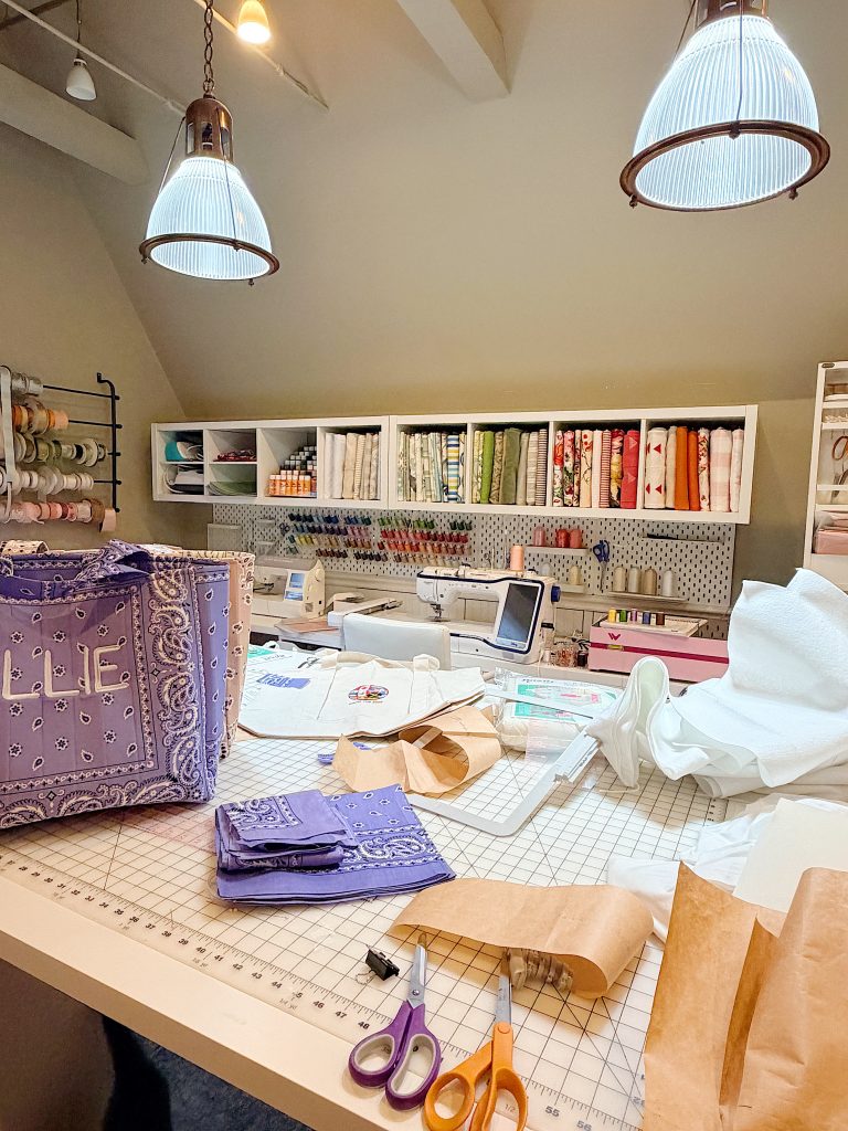 A sewing workspace with fabric, scissors, paper patterns, and sewing machines on a cutting table, with shelves of fabric and supplies in the background.