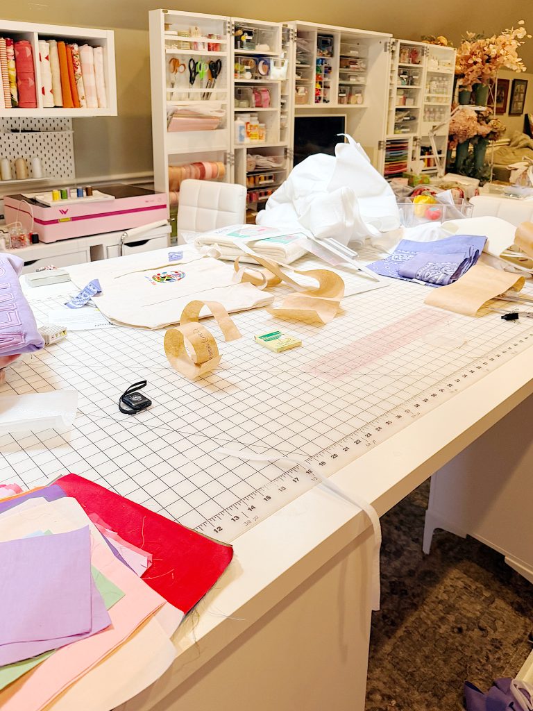 A craft room workspace with a large cutting mat, fabric pieces, measuring tools, and various craft supplies scattered on a table, with storage shelves in the background.