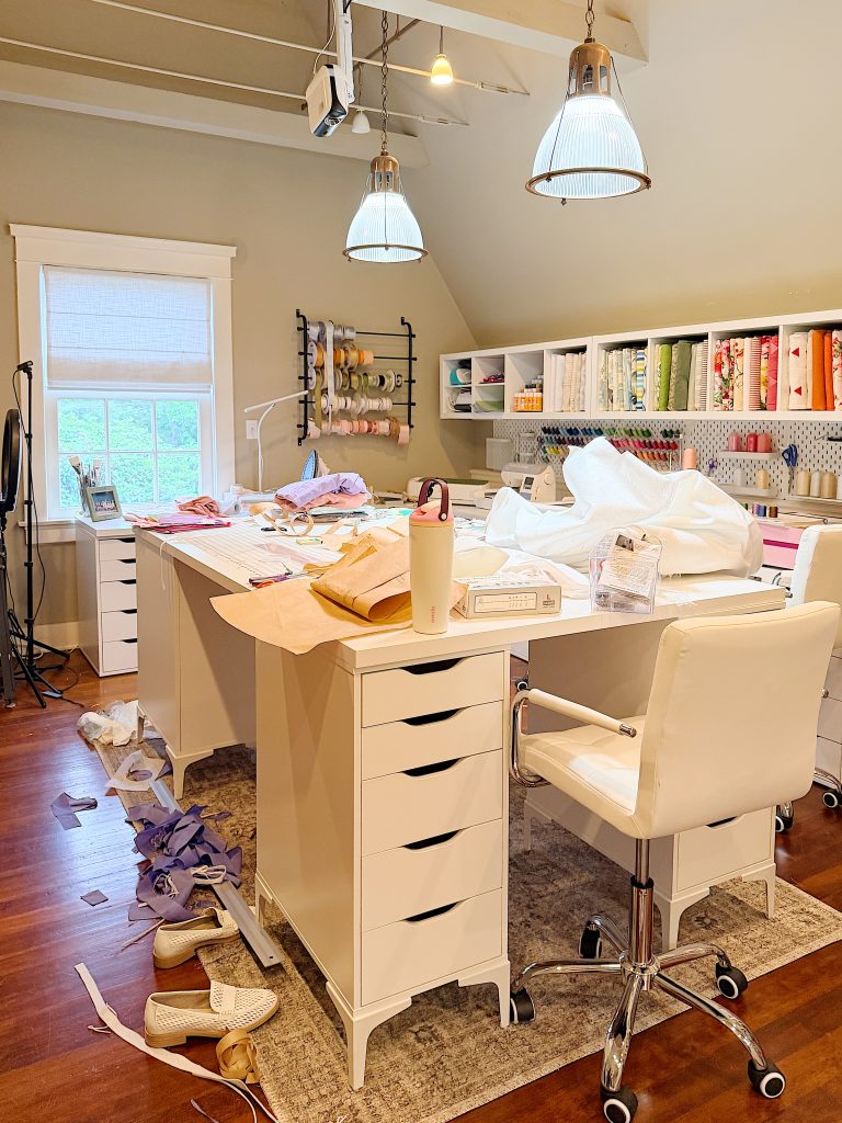 A craft room with a white desk cluttered with fabrics and sewing tools, shelves with organized supplies, a chair, and shoes on the floor.