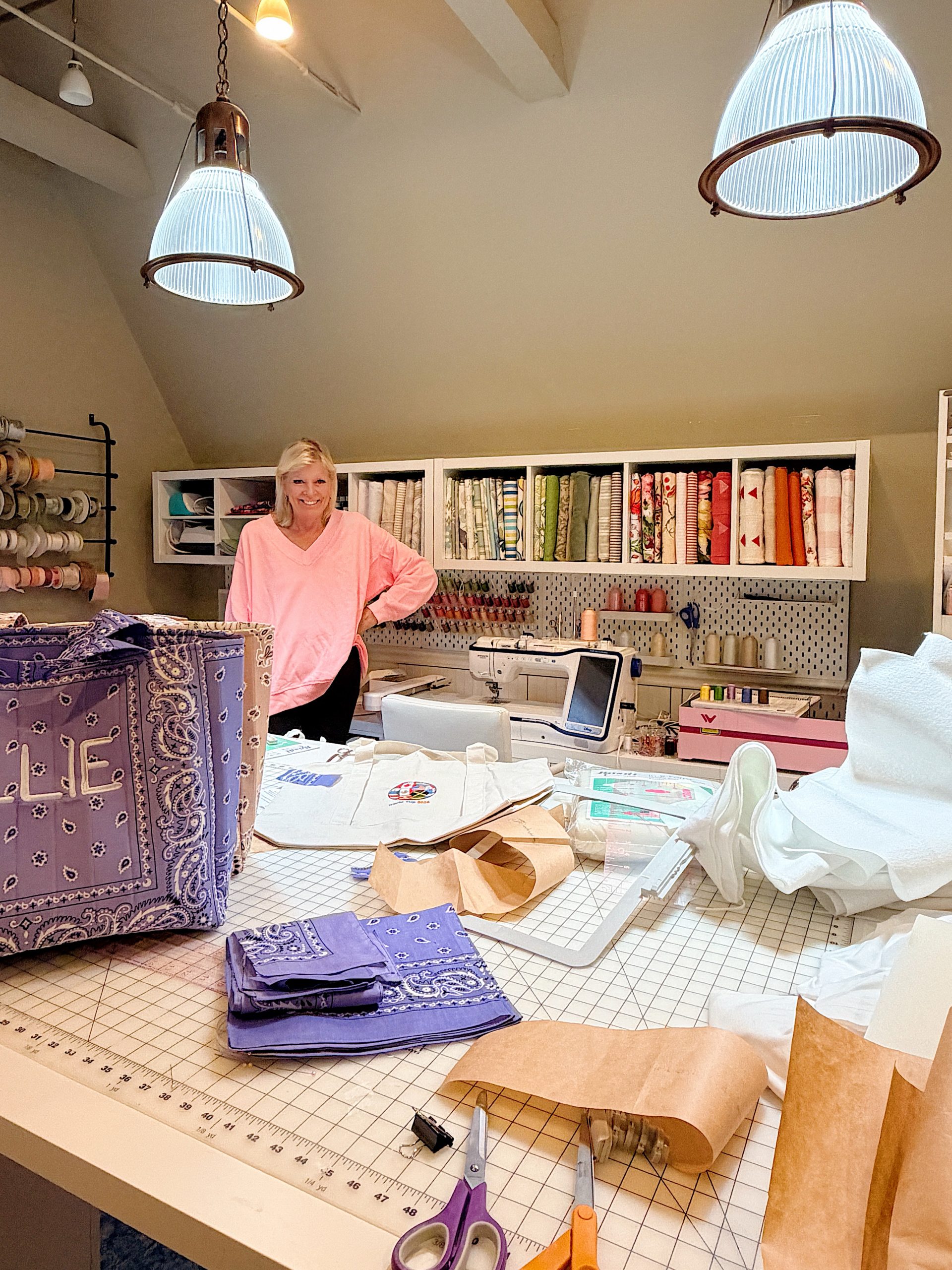 A woman stands in a sewing room with fabric, patterns, and tools spread on a worktable. Shelves with fabric and sewing supplies are in the background.