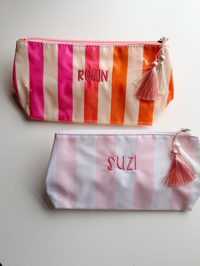 Two striped zipper pouches with tassels on a white surface; the top pouch reads "ROBIN" in pink and orange, the bottom reads "SUZI" in pink and white.