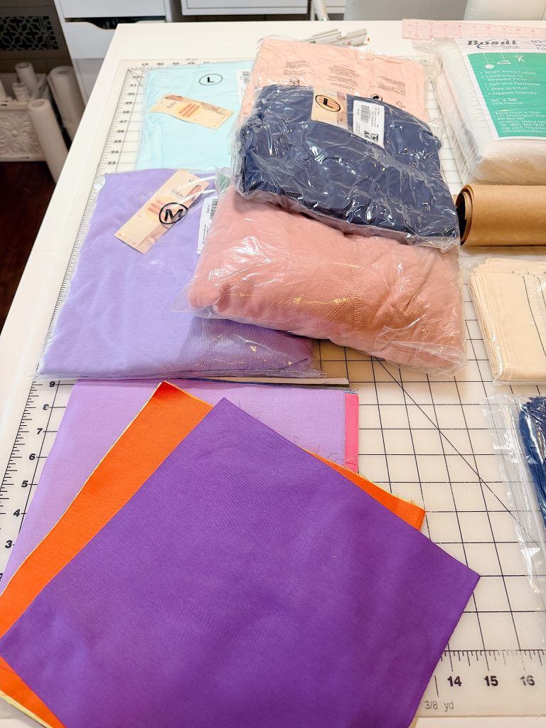 Stacks of folded fabric, some in plastic packaging, sit on a cutting mat along with loose fabric squares in purple and orange colors.