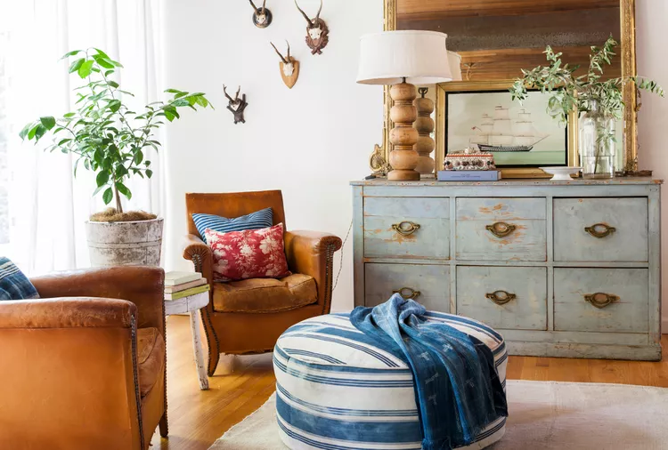 Living room with two brown leather chairs, a striped ottoman, blue chest of drawers, potted plant, lamp, framed art, and antler wall decor.