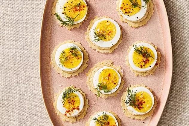 A pink oval plate holds eight crackers topped with slices of hard-boiled egg, a dollop of spread, dill sprigs, and a sprinkle of seasoning, arranged neatly on a beige tablecloth.