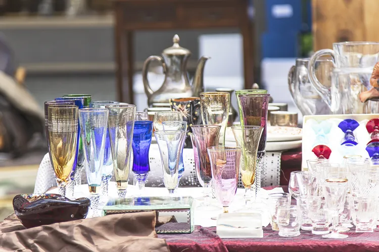 A variety of colorful vintage glassware, cups, and a metal teapot are displayed on a table at an outdoor market.