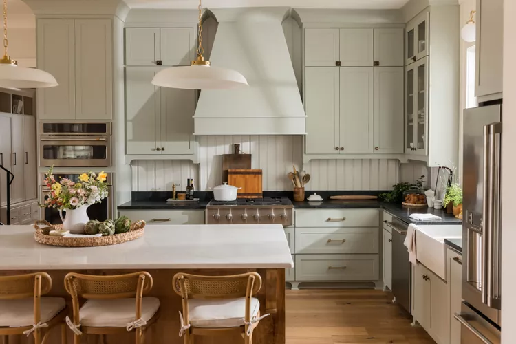 Modern kitchen with light green cabinets, central island with white countertop and wicker chairs, stainless steel appliances, and decorative pendant lights overhead.