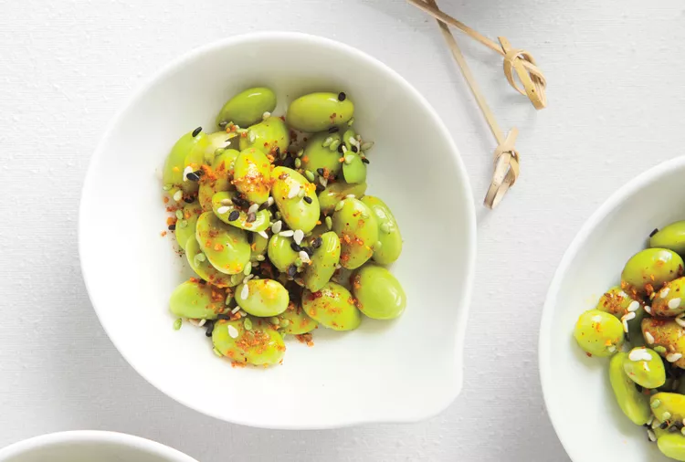A white bowl filled with seasoned edamame beans topped with sesame seeds and spices, placed on a white surface.