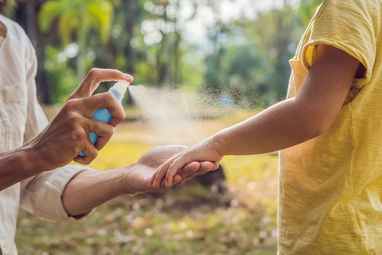 An adult sprays insect repellent onto a child’s arm outdoors, with trees and grass visible in the background.