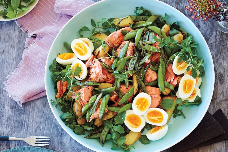 A salad with soft-boiled eggs, green beans, potatoes, sugar snap peas, salmon pieces, and greens in a large bowl on a wooden table.