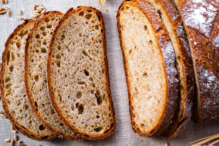 Four slices of rustic brown bread are arranged on a linen surface next to an unsliced portion of the loaf.