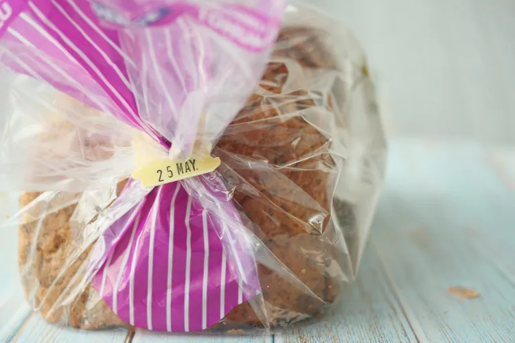 A plastic bag of sliced bread with a twist tie showing an expiration date of 25 May, placed on a light wooden surface.
