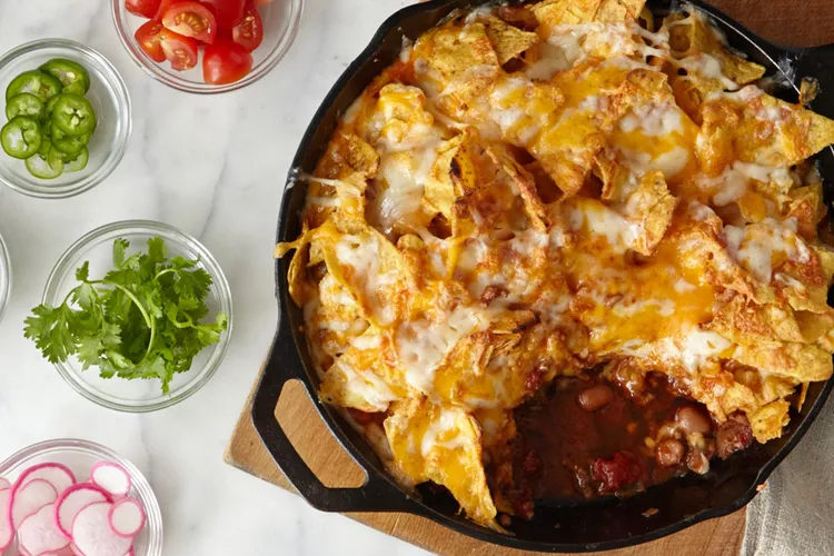A cast iron skillet filled with cheesy nachos and beans, surrounded by bowls of sliced jalapeños, cherry tomatoes, cilantro, and radishes on a marble surface.