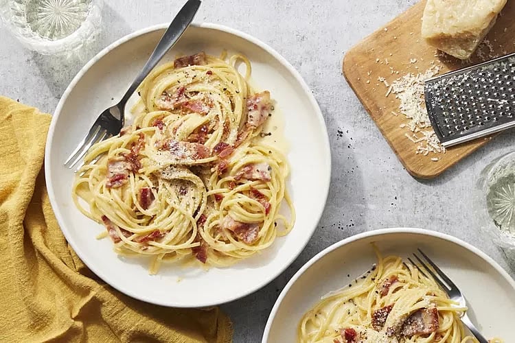 Two plates of spaghetti carbonara with bacon, served with forks. A cheese grater and a block of parmesan cheese are on a cutting board nearby. A yellow napkin and glasses of water are also present.