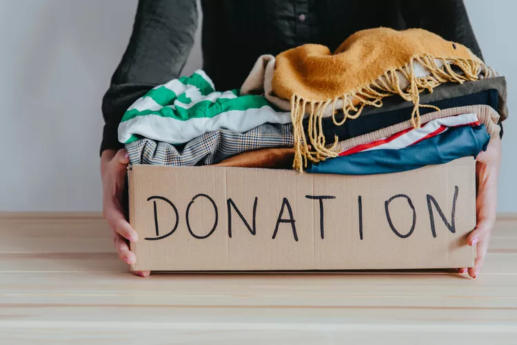 Person holding a cardboard box labeled "DONATION" filled with assorted folded clothes, positioned on a wooden table.