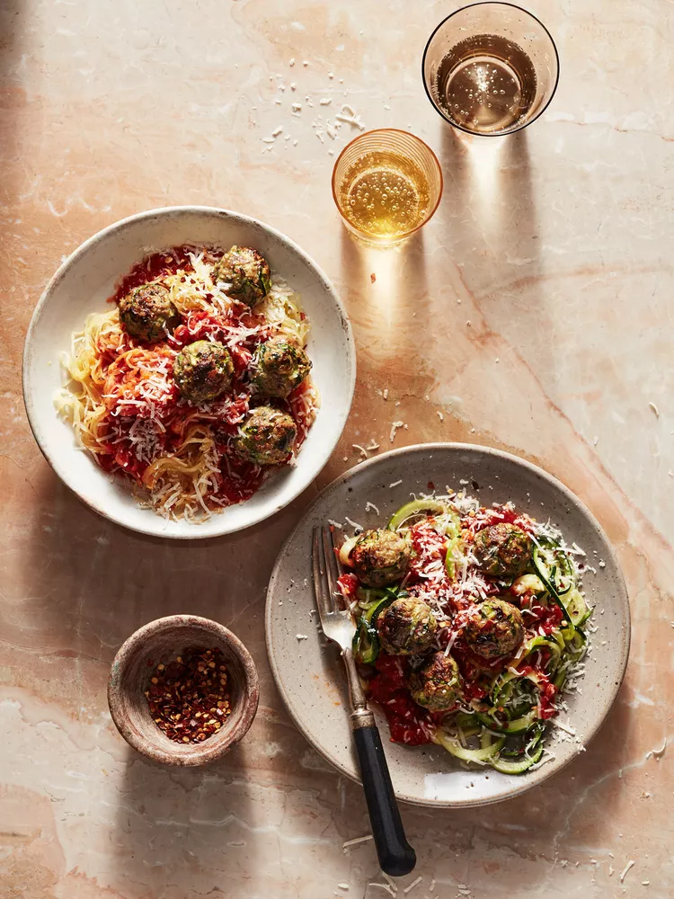 Two plates of vegetable noodles with tomato sauce, veggie meatballs, and grated cheese, accompanied by two glasses of beverage and a bowl of red pepper flakes on a marble surface.